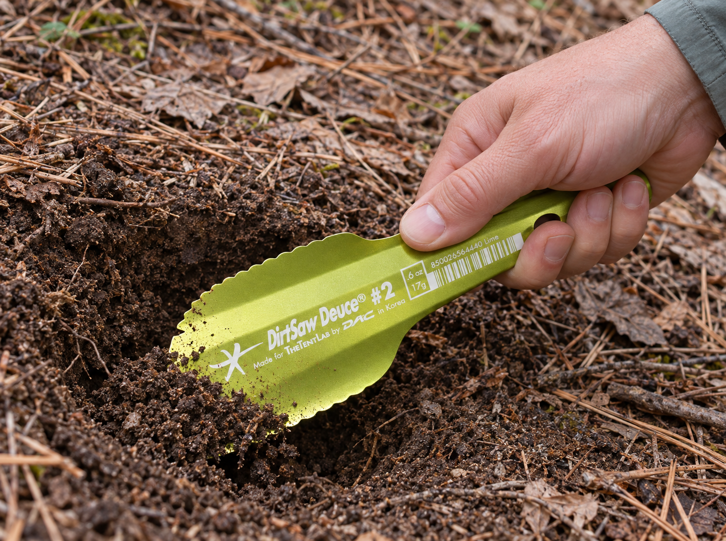Hand gripping a lime DirtSaw Deuce #2 trowel while scooping loose soil at a small forest-floor hole, showing its curved blade profile