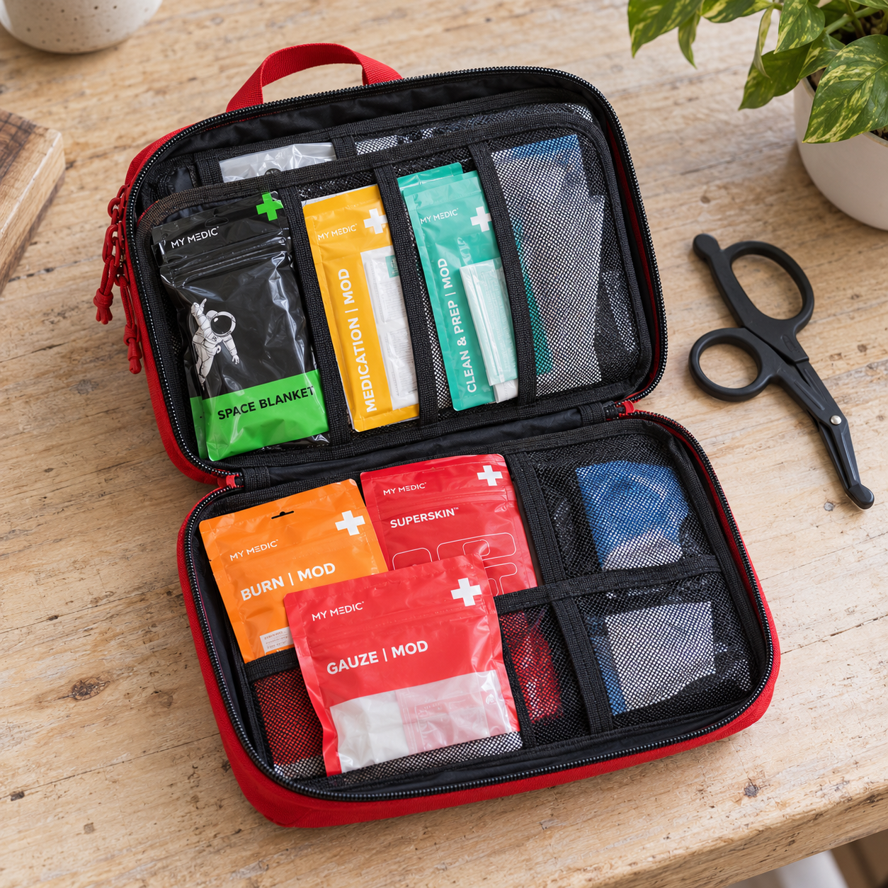 Open red MyMedic MyFAK Standard kit with color-coded supply modules in black mesh organizers and shears on a wooden table