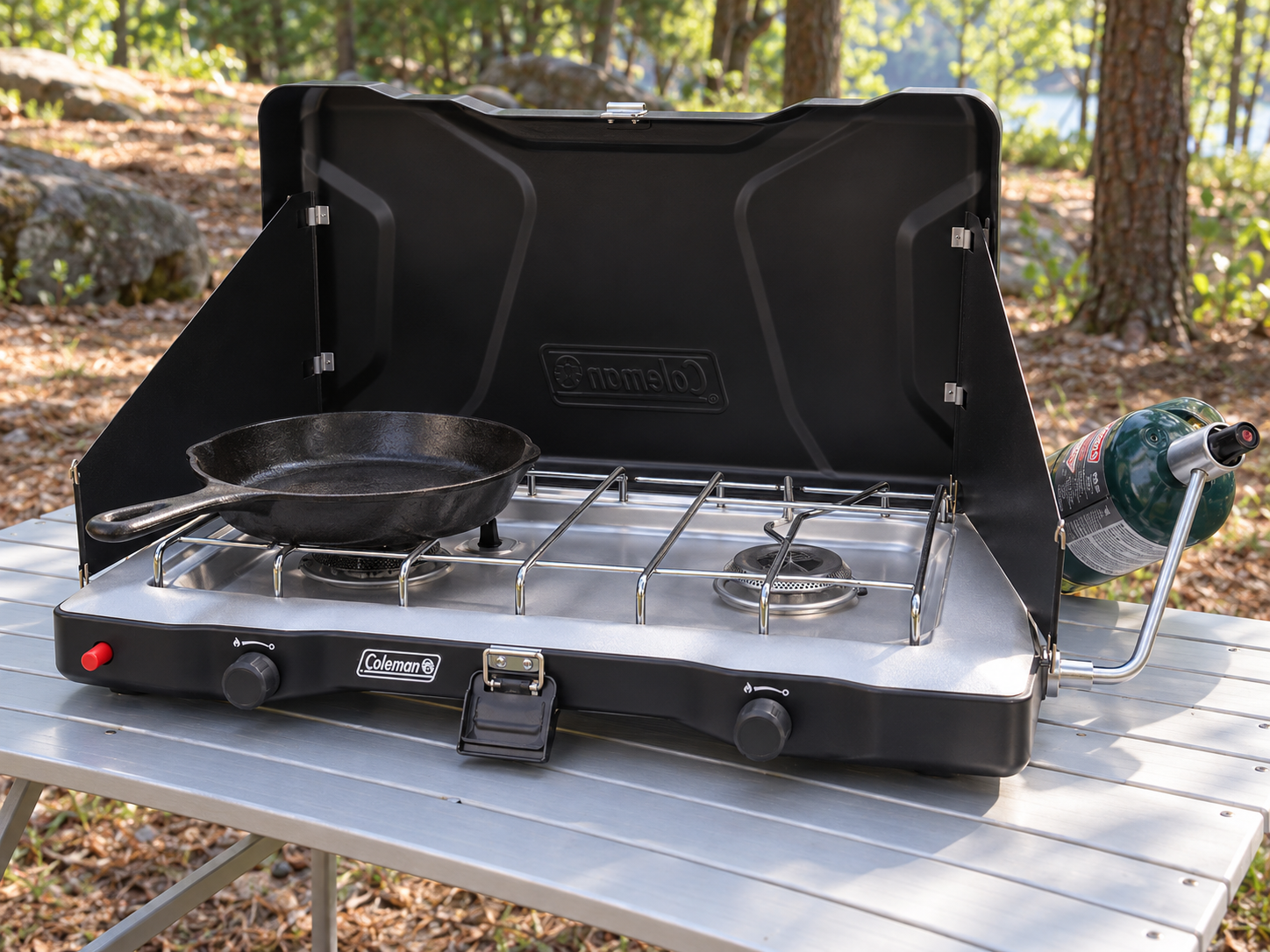 Open black Coleman Triton+ two-burner stove with a skillet on an outdoor camp table and a connected propane cylinder beside it.