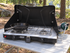 Open black Coleman Triton+ two-burner stove with a skillet on an outdoor camp table and a connected propane cylinder beside it.