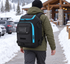 Adult wearing the black ski boot backpack with blue trim on both shoulders while walking along a snowy ski-lodge path