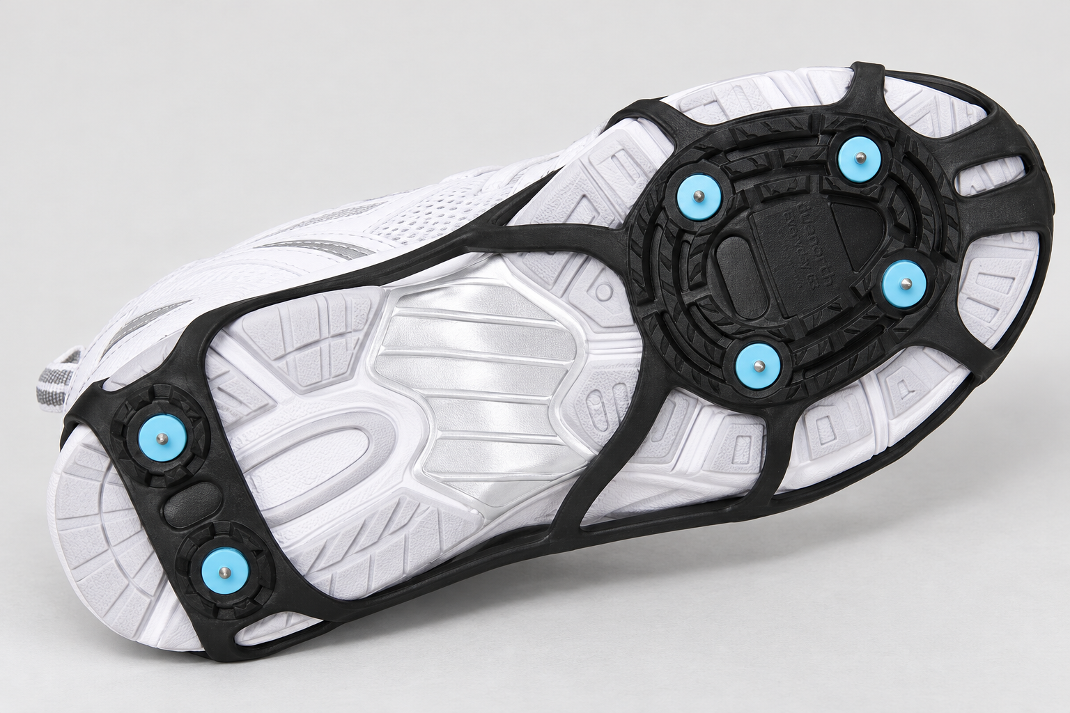 Underside of a black Due North Everyday G3 traction aid on a white athletic shoe, showing four forefoot spikes and two heel spikes.
