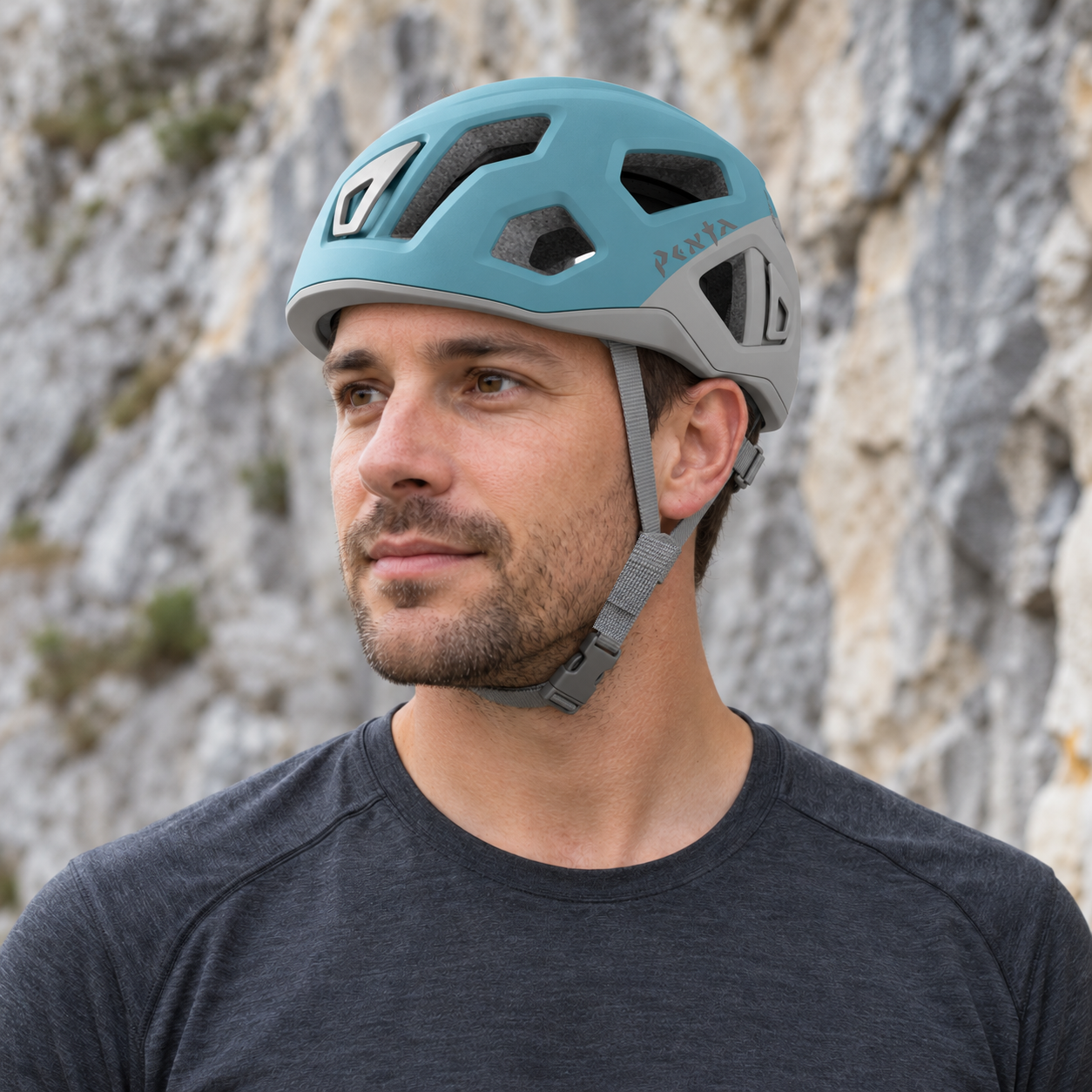 Adult in three-quarter view wearing a Spruce Blue Singing Rock Penta 2 helmet at a rocky crag, showing grey ear-side straps, a woven below-ear sleeve and chin buckle.