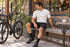 Cyclist in black DAREVIE DVP008 bib shorts seated on a coffee-shop bench with his bicycle nearby and a cup on the side table
