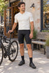 Cyclist wearing black DAREVIE DVP008 bib shorts and an ivory jersey beside a bicycle outside a neighborhood bike shop