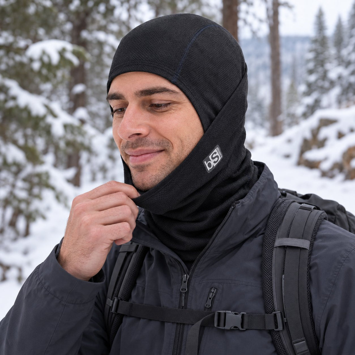 An adult hiker touches the lowered face panel of the BlackStrap Merino Hood in Black on a snowy forest trail.