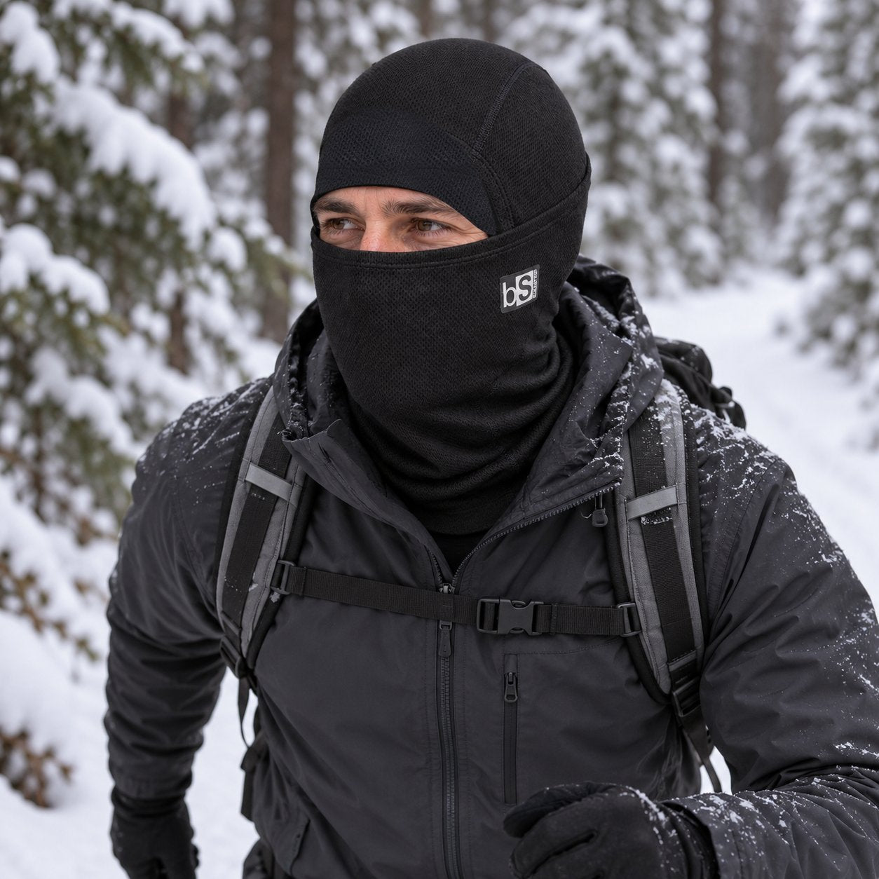 An adult hiker wears the BlackStrap Merino Hood in Black with the face panel raised on a snowy forest trail.
