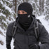 An adult hiker wears the BlackStrap Merino Hood in Black with the face panel raised on a snowy forest trail.