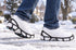 Black Due North Everyday G3 traction aids fitted to white athletic shoes during a walking step on a snow-covered neighborhood path.