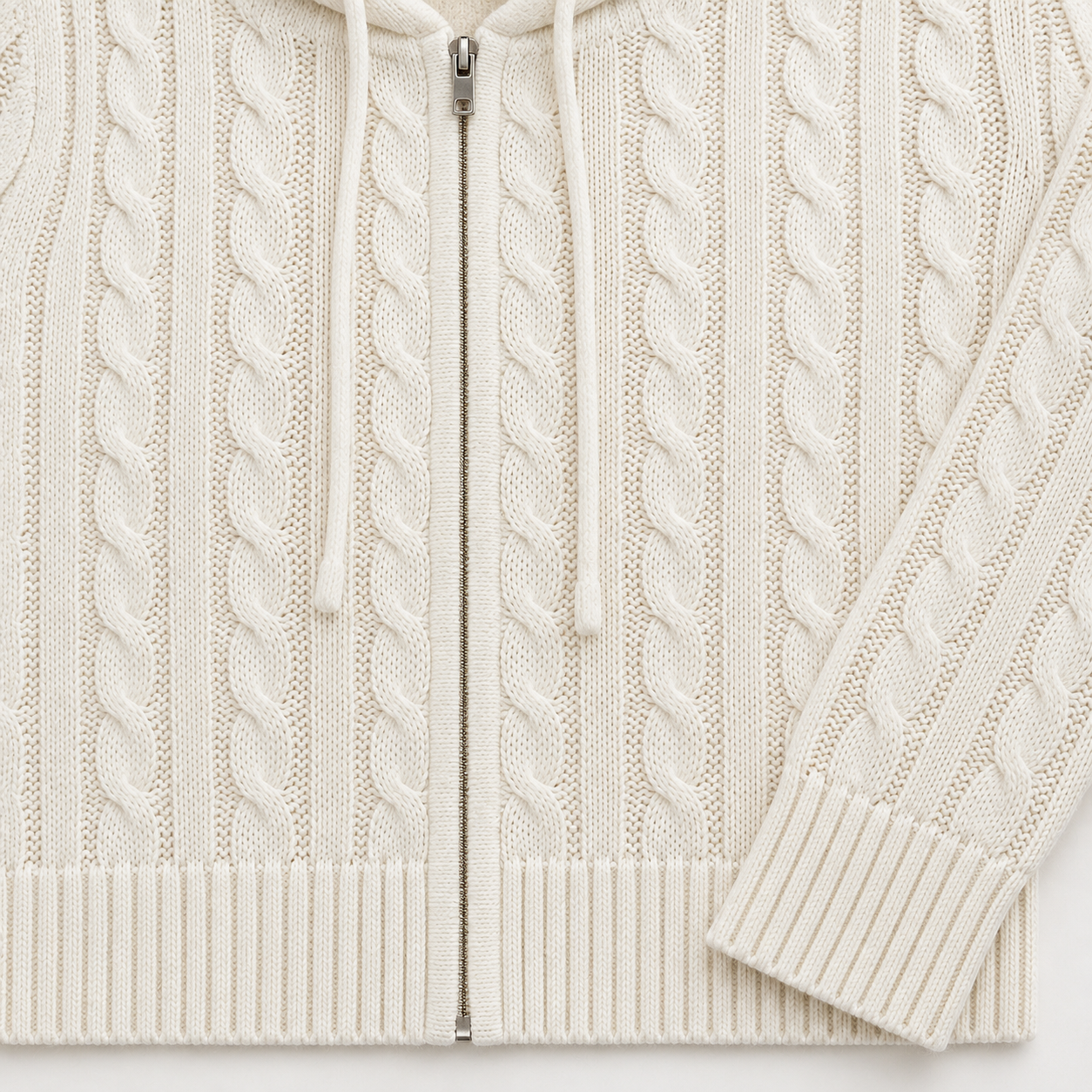 Close view of the cream cable-knit hoodie front showing the central zip, cream drawstrings, vertical cable pattern, ribbed hem and ribbed cuff.