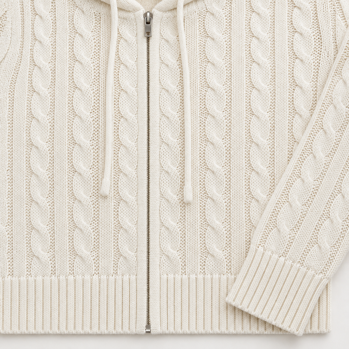 Close view of the cream cable-knit hoodie front showing the central zip, cream drawstrings, vertical cable pattern, ribbed hem and ribbed cuff.