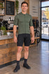 Man wearing an olive YKYWBIKE jersey and fitted black cycling shorts in a coffee shop.