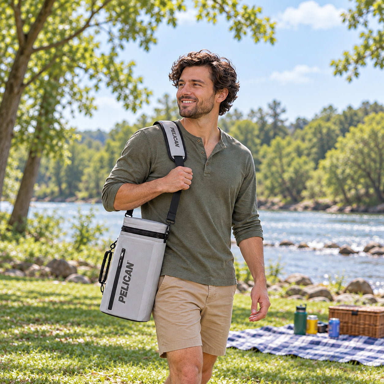 A man carrying a gray Pelican Dayventure Sling Cooler to a riverside park picnic.